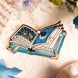 May include: A detailed enamel pin in the shape of an open book. The book has blue and gold accents, with the words "MOON-BREW" on the left page. The right page features a crescent moon and stars. The pin is a whimsical accessory.