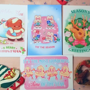 May include: Set of four Christmas-themed greeting cards featuring illustrations of a bear, bunnies, a reindeer, and bells. The cards are printed on white cardstock with colorful illustrations and text.