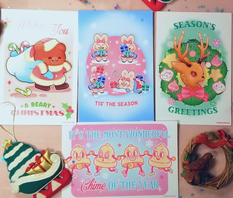 May include: Set of four Christmas-themed greeting cards featuring illustrations of a bear, bunnies, a reindeer, and bells. The cards are printed on white cardstock with colorful illustrations and text.