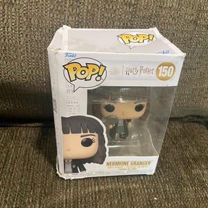 May include: A Funko Pop! vinyl figure of Hermione Granger from the Harry Potter series. The figure is wearing her Hogwarts uniform and holding a Time-Turner. The box is white with the Funko Pop! logo and the Harry Potter logo. The number 150 is printed on the box.