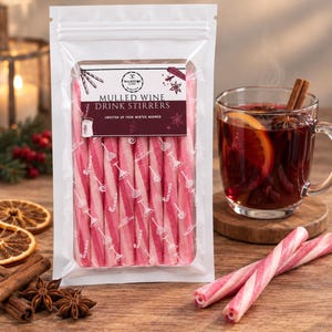 May include: A clear plastic pouch containing pink and white striped cylindrical candy sticks labeled Mulled Wine Drink Stirrers. The label features a logo, text, and small illustrations of candy. Two loose candy sticks lie on the surface nearby.