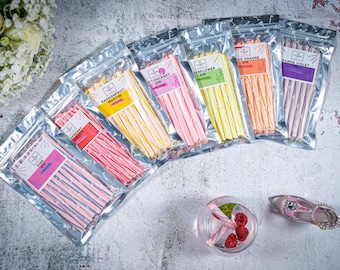 Op de afbeelding: Assortiment van kleurrijke snoepstokjes in individuele doorzichtige plastic zakjes. Elk zakje heeft een label met een andere smaak en gin-naam, zoals Raspberry Martini, Cotton Candy Gin en Wood Orange Gin.