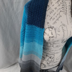 K&ouml;nnte beinhalten: Eine gestrickte Strickjacke in Blau, Grau und Wei&szlig; mit einem langen, offenen Vorderteil.