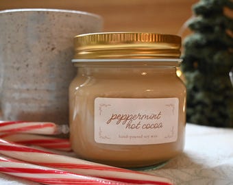 May include: A jar of peppermint hot cocoa scented soy wax candle with a gold lid. The label is white with brown text. Candy canes and a ceramic mug are in the background.