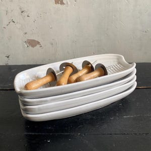 May include: Four white ceramic serving dishes stacked, each with a ridged design. Each dish holds corn cob holders with wooden handles and metal prongs. The dishes are on a dark surface.