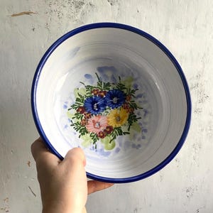 May include: White ceramic bowl with a blue rim and floral design. The bowl features a hand-painted bouquet of blue, pink, and yellow flowers with green leaves. The bowl is held by a person's hand.