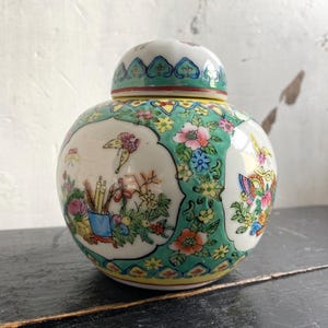 May include: A round, lidded ceramic jar with a floral and butterfly design. The jar has a white base with a green border and is decorated with colourful flowers and butterflies. The lid is white with a green and blue pattern.