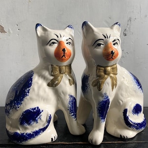 May include: Two ceramic cat ornaments. The cats are white with blue markings, orange noses, and gold bow ties. They are depicted in a seated position with curled tails. The ornaments have a vintage appearance.