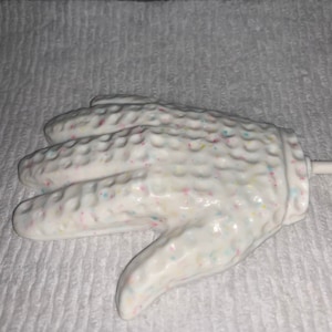 May include: A white candy lollipop shaped like a hand with colorful sprinkles. The lollipop is on a white surface.