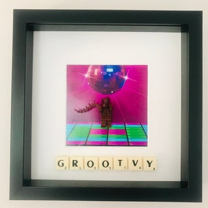 May include: A framed image of a brown Lego figure standing on a dance floor with a disco ball above. The figure is surrounded by colourful tiles. The word "GROOVY" is spelled out below the image using letter tiles.
