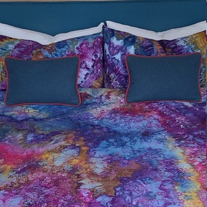 Può includere: Un letto con una testiera blu-verde e un copripiumino astratto colorato. Due cuscini blu scuro con bordatura rossa sono sul letto.