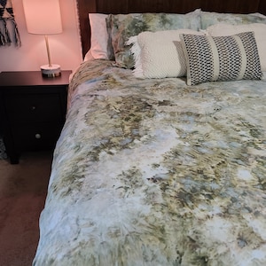 Pode incluir: Uma cama com uma capa de edredom branca com um padrão abstrato verde e azul. A cama é feita com almofadas brancas e creme, incluindo uma com um padrão geométrico preto e branco.