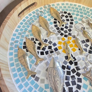 Peut inclure: Une collection de porte-clés métalliques en forme de poisson disposés sur un plateau circulaire. Plusieurs porte-clés sont en laiton avec des motifs d'écailles gravés, tandis qu'un plus grand porte-clé central est argenté avec des gravures décoratives.