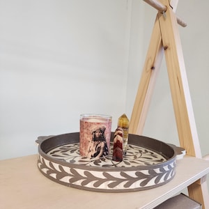 Op de afbeelding: Een ronde dienblad met een grijs en wit bloemenmotief. Het dienblad heeft een verhoogde rand en twee handvatten. Een glazen kaarsenhouder met een bloemenmotief en een vrouwengezicht staat op het dienblad. Twee kristallen punten staan ook op het dienblad.