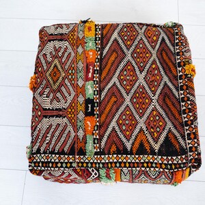 May include: Square, patterned floor cushion in shades of red, orange, black, and cream. The cushion features geometric diamond and stripe designs, with colorful tassels on the corners. The cushion measures approximately 61 cm square.