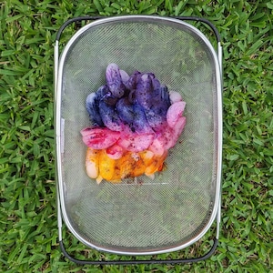 May include: A metal mesh colander filled with colorful, dyed fabric. The fabric is arranged in layers of orange, pink, purple, and blue, creating a gradient effect. The colander sits on a bed of green grass.