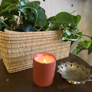 Pode incluir: Uma vela laranja-avermelhada acesa em um suporte cilíndrico está em uma superfície de madeira escura ao lado de uma tigela decorativa de metal. Uma cesta de vime cheia de plantas verdes folhosas está no fundo, criando um tema de decoração natural.