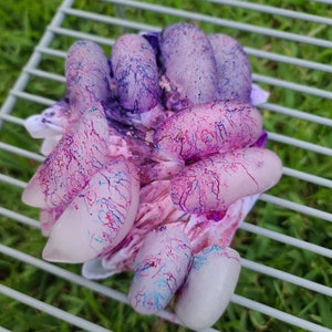 May include: A collection of painted fingernails in various shapes and sizes, showcasing a marbled design with shades of pink, purple, and blue. The nails are arranged on a white cloth, resting on a metal rack, with a blurred green background.