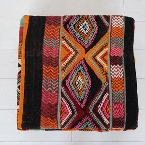 May include: Square, patterned ottoman with a black background and colourful geometric designs in orange, pink, and teal. The woven fabric features diamond and zigzag patterns. A decorative accent for a living room or bedroom.