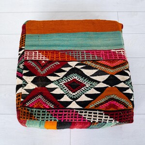 Puede incluir: Otomana cuadrada y estampada con una vibrante mezcla de colores. La parte superior presenta bandas naranjas y turquesas, mientras que los lados muestran diseños geométricos en rojo, negro, blanco y rosa. Esta pieza decorativa añade un toque bohemio a cualquier espacio.