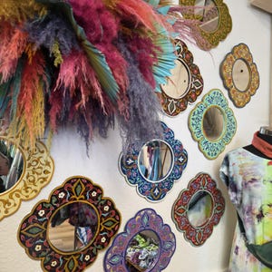 May include: A collection of decorative wall mirrors with ornate, colorful frames. The mirrors are in various shapes and sizes, with intricate patterns in shades of blue, red, green, and gold. A vibrant arrangement of dried flowers in pink, purple, and yellow adds a pop of color.