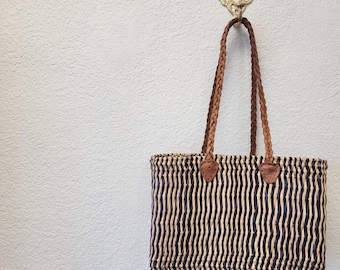 May include: A rectangular tote bag with vertical black and beige stripes. The bag has braided brown leather handles and leather accents. It hangs from a decorative gold-colored wall hook.