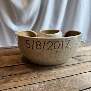 May include: Speckled beige ceramic serving bowl with three sections and a central, smaller bowl. The date "5/8/2017" is etched on the front. The bowl sits on a rustic wooden surface.