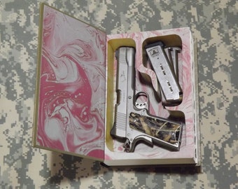 May include: A silver handgun with a camouflage grip is stored inside a book safe with a pink and white marbled design. Two silver magazines are also stored in the safe.