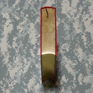 May include: A hardback book with a red spine and gold-coloured pages. A black bookmark hangs from the top. The book is standing upright against a camouflage background.