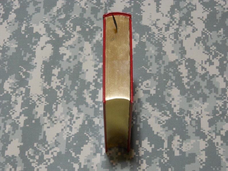 May include: A hardback book with a red spine and gold-coloured pages. A black bookmark hangs from the top. The book is standing upright against a camouflage background.