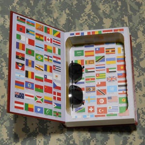 May include: A decorative book with a hollowed-out interior, featuring a collection of international flags on the inside pages. Black sunglasses rest inside the book. The book's cover is red, and the background is camouflage.