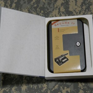 May include: A black Secure It handgun safe, designed to look like a book, is open to reveal the safe. The safe has a key lock and is nestled in a white foam insert. The book's pages are white.
