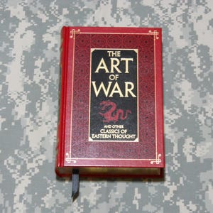 May include: A red hardback book titled "THE ART OF WAR" with gold lettering. The cover has a black panel with a red dragon illustration. A black ribbon bookmark is visible. The book is a classic of Eastern thought.