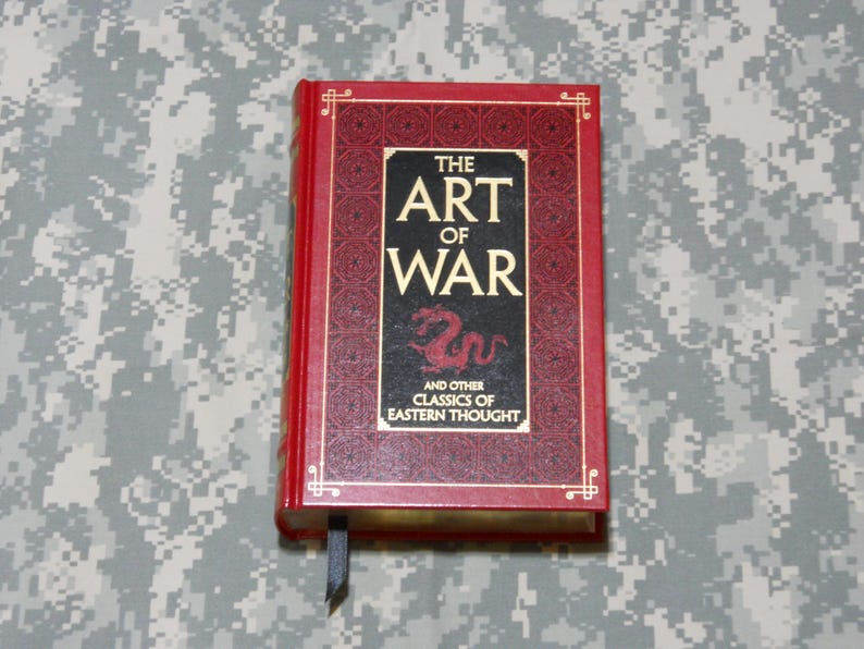 May include: A red hardback book titled "THE ART OF WAR" with gold lettering. The cover has a black panel with a red dragon illustration. A black ribbon bookmark is visible. The book is a classic of Eastern thought.