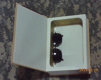 May include: A white book with a hidden compartment that opens to reveal a pair of black sunglasses. The book is lying flat on a camouflage surface.