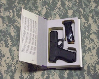 May include: An open book with a hidden compartment containing a black handgun and a magazine. The book is on a camouflage patterned surface. The pages of the book are visible.