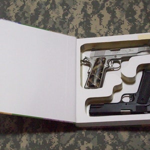 May include: Two handguns are hidden inside a book. The book is open and the guns are in their cutouts. The top gun has a camouflage grip and the bottom gun has a black grip.