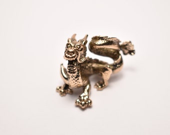 May include: A gold-colored dragon figurine with a curled tail and detailed scales. The dragon is facing forward with its mouth open.