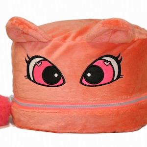 Op de afbeelding: Een koraalkleurige opbergdoos van pluche met een cartoongezicht met grote roze ogen, zwarte pupillen en kleine hartjes. De doos heeft twee kleine oortjes aan de bovenkant, een horizontale ritssluiting en een ronde, felroze pompon als ritstrekker.