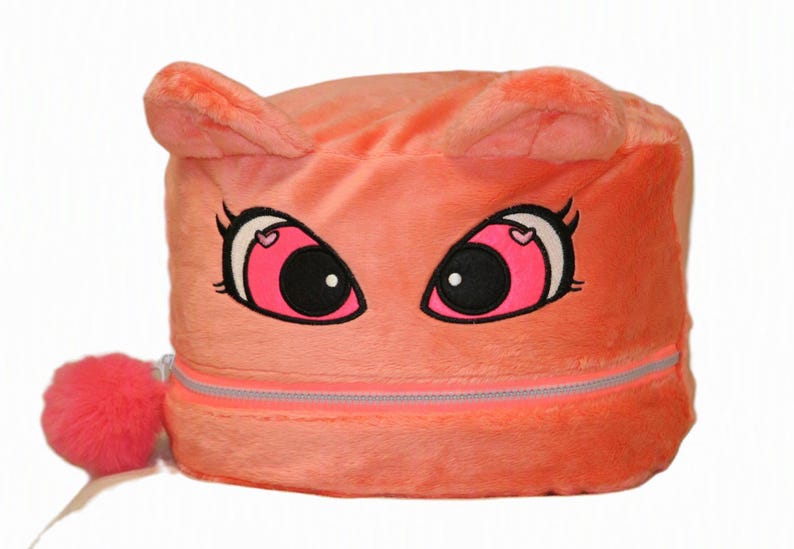Op de afbeelding: Een koraalkleurige opbergdoos van pluche met een cartoongezicht met grote roze ogen, zwarte pupillen en kleine hartjes. De doos heeft twee kleine oortjes aan de bovenkant, een horizontale ritssluiting en een ronde, felroze pompon als ritstrekker.