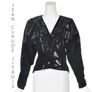 Peut inclure: Veste en daim noir avec un motif géométrique en cuir noir. La veste a un devant enroulé et des manches longues.