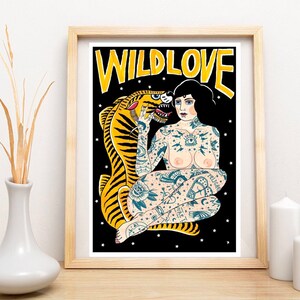 May include: A framed print with a black background featuring a woman with tattoos sitting with a tiger. The woman has black hair and is wearing a necklace. The tiger is yellow and black. The text "WILDLOVE" is written in yellow letters at the top of the print.