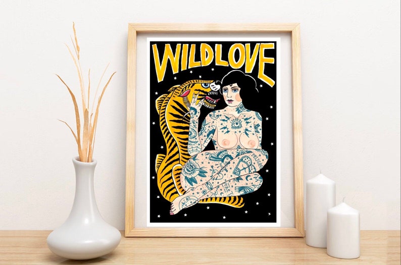 May include: A framed print with a black background featuring a woman with tattoos sitting with a tiger. The woman has black hair and is wearing a necklace. The tiger is yellow and black. The text "WILDLOVE" is written in yellow letters at the top of the print.