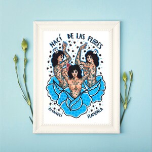 Puede incluir: Una impresión enmarcada de una ilustración colorida de tres mujeres con tatuajes, de pie en una flor azul. El texto "NACI DE LAS FLORES AMANECI FLAMENCA" está escrito alrededor de las mujeres.