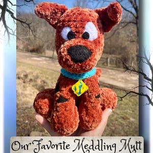 May include: A brown plush dog toy made of soft yarn with large white and black eyes, a black nose, and a bright blue collar featuring a yellow diamond-shaped tag with blue details. The text reads Our Favorite Meddling Mutt Crochet Pattern.