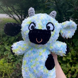 Pode incluir: Uma vaca de pelúcia de crochê com um padrão de bolinhas azuis e verdes. A vaca tem orelhas pretas, um nariz preto e uma mancha preta no lado. A vaca tem um anel no nariz.