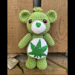 Peut inclure: Ours en crochet vert avec des yeux rouges et une poitrine blanche ornée d'une feuille de marijuana verte.
