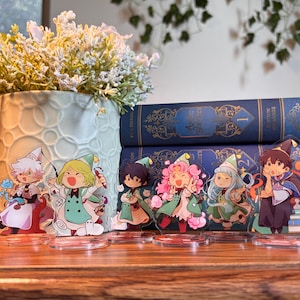 May include: Six individual acrylic character standees featuring chibi-style figures in colorful fantasy outfits. Each figure is mounted on a clear circular base, displaying distinct poses and expressions, arranged in a row on a wooden surface.