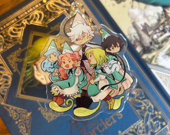 May include: A clear acrylic charm featuring a group of five illustrated characters wearing pointed hats and teal capes. The central figure has white hair and round glasses, surrounded by four others with varied hair colors, including pink and green.