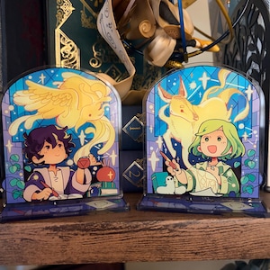 May include: Two acrylic bookends with colorful illustrations. One features a person writing with a quill, a stack of books, and a yellow winged creature. The other shows a person with green hair, a yellow creature, and a white bird.